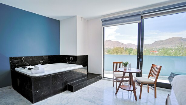 A modern bathroom with a black marble bathtub, a small table with chairs, and a large window with a view of mountains. - Hotel Guiant (Santa María del Río)