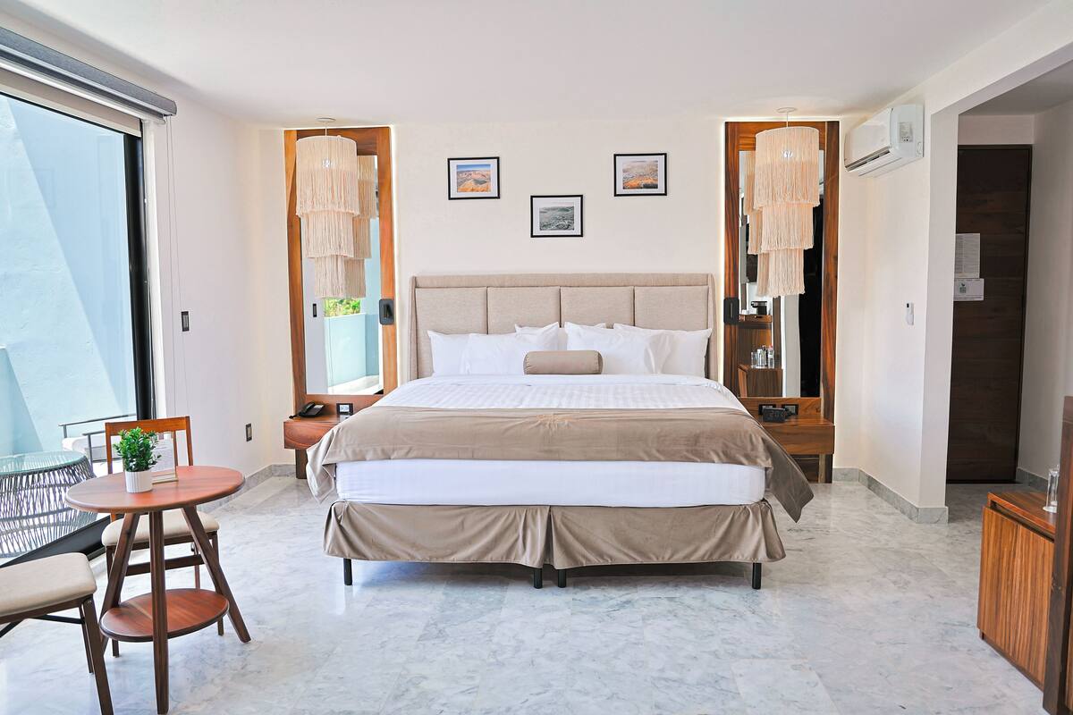 A hotel room with a large bed, a small round table with a chair, and a balcony with a glass railing.