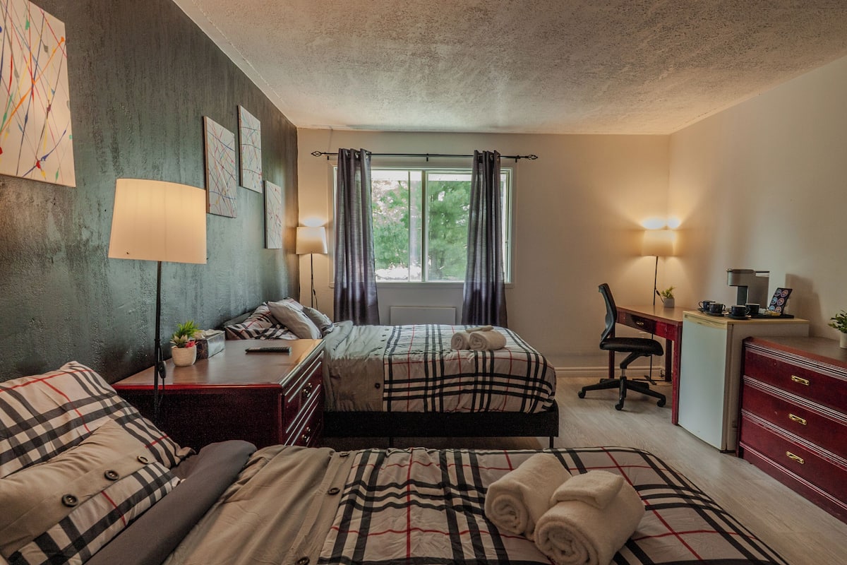 A bedroom with two beds, a desk, a chair, and a dresser. There are curtains on the window, and a lamp on the desk.