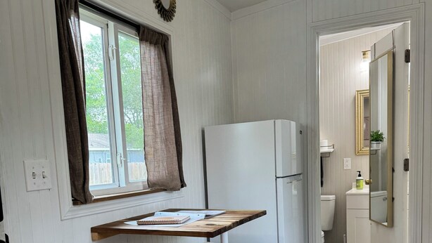 A compact kitchen with a white refrigerator, a wooden table, and two white stools. A window with curtains is visible, and a ceiling fan is installed. - Sunnyside Suit, Luxe Tiny Home Gulfport, MS (Gulfport)