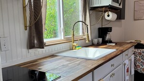A kitchen with a wooden countertop, white cabinets, a sink, a microwave, and a window with a view of greenery. - Sunnyside Suit, Luxe Tiny Home Gulfport, MS (Gulfport)