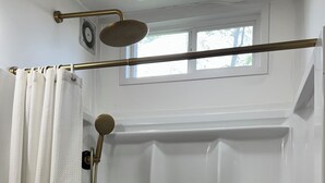 A shower with a handheld showerhead, a window above, and shelves with toiletries. - Sunnyside Suit, Luxe Tiny Home Gulfport, MS (Gulfport)