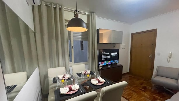 Un comedor con una mesa dispuesta para cuatro personas, un televisor sobre un mueble bajo y un sofá al fondo.