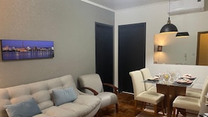 Una sala de estar con un sofá, sillas, una mesa de comedor preparada para dos personas y un cuadro de paisaje urbano en la pared.