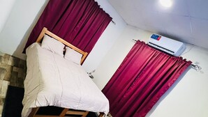 Quarto com cama, duas janelas com cortinas vermelhas, ar condicionado, uma pequena planta e piso de madeira.