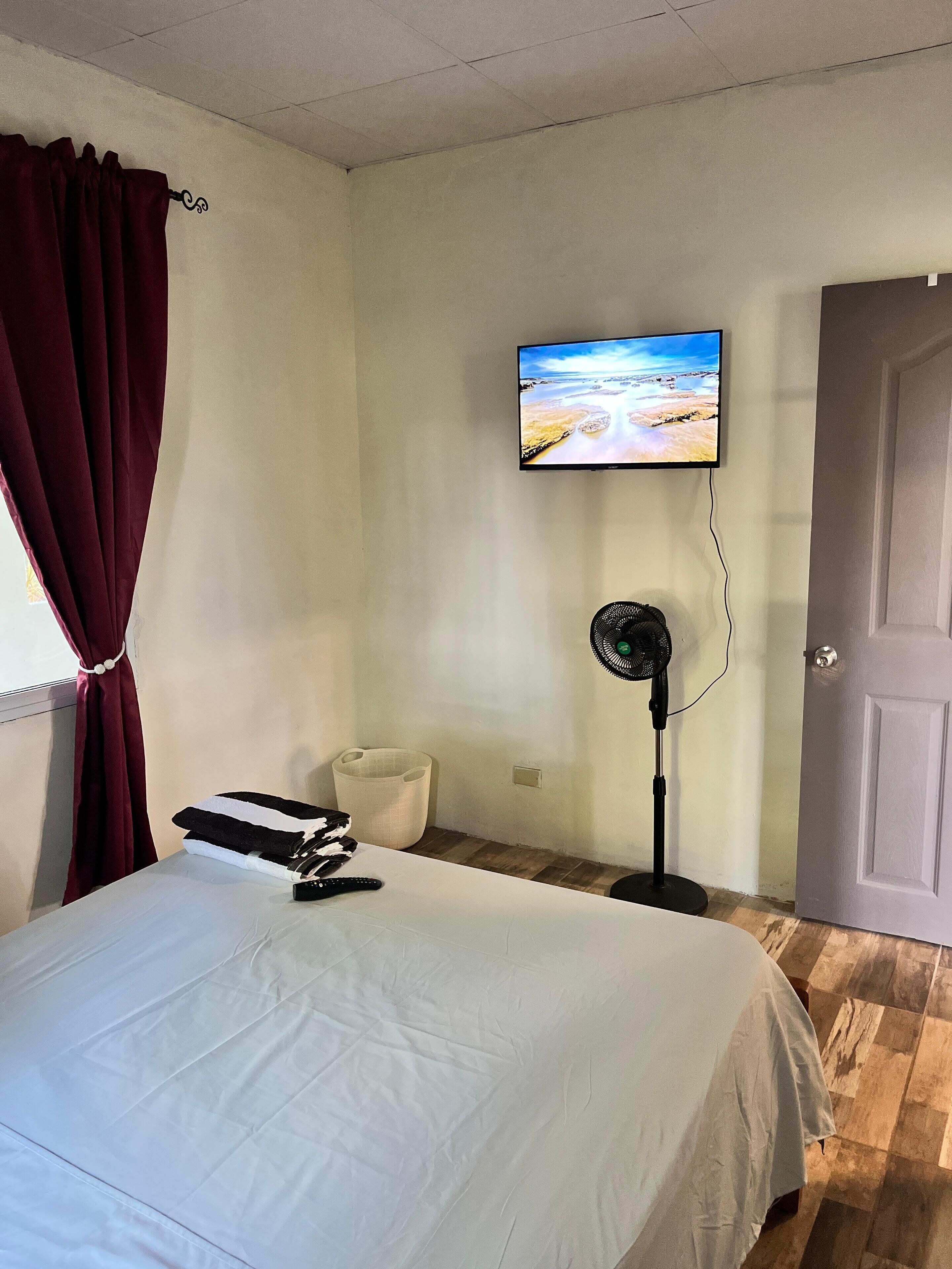 Une chambre d’hôtel avec un lit, un téléviseur fixé au mur, un ventilateur sur pied et une porte.
