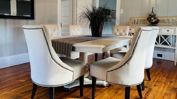 Een eetgedeelte met een witte tafel, vier gestoffeerde stoelen met zwarte accenten, een kroonluchter en een grote plant.