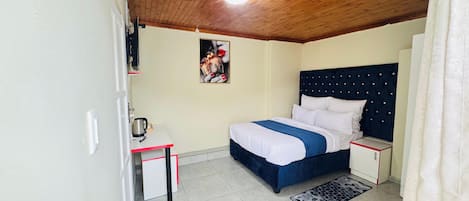 Un dormitorio con techo de madera, una cama con cabecera azul, una mesita de noche y una alfombra de bienvenida.