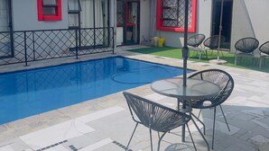 Una piscina con un borde de baldosas, un porche cubierto con mobiliario exterior y un edificio con ventana de marco rojo y el número 118.