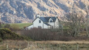 A house with a gabled roof, surrounded by a fence and vegetation, with mountains in the background. - Cuillin View House (Arisaig)