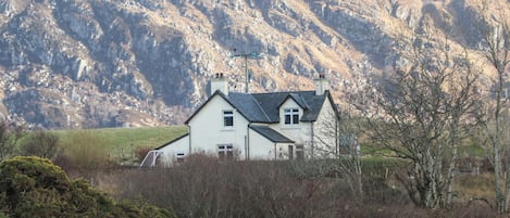Ein Haus mit Satteldach, umgeben von einem Zaun und Vegetation, mit Bergen im Hintergrund.