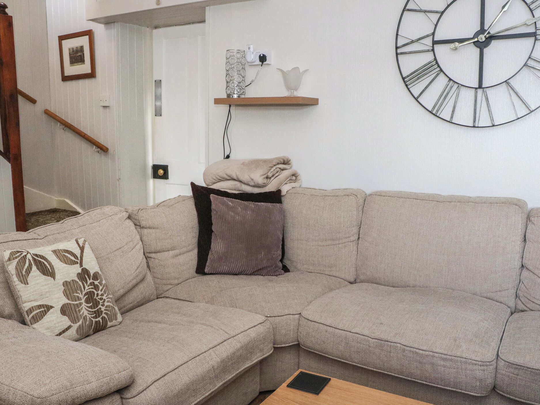 A living room with a sectional sofa, a round wall clock, a staircase, and a small shelf with decorative items.