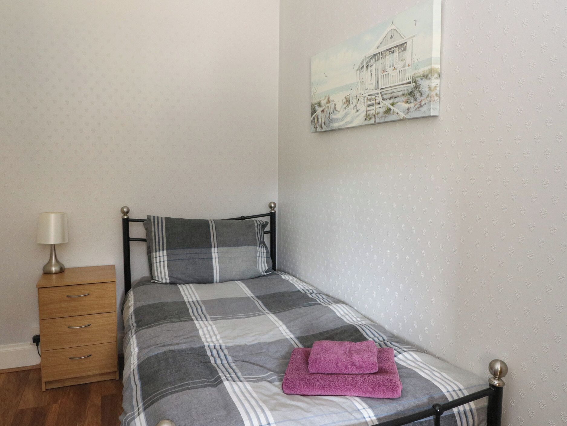 A single bed with a checkered bedspread, a pillow, a wooden nightstand with a lamp, and a painting on the wall.