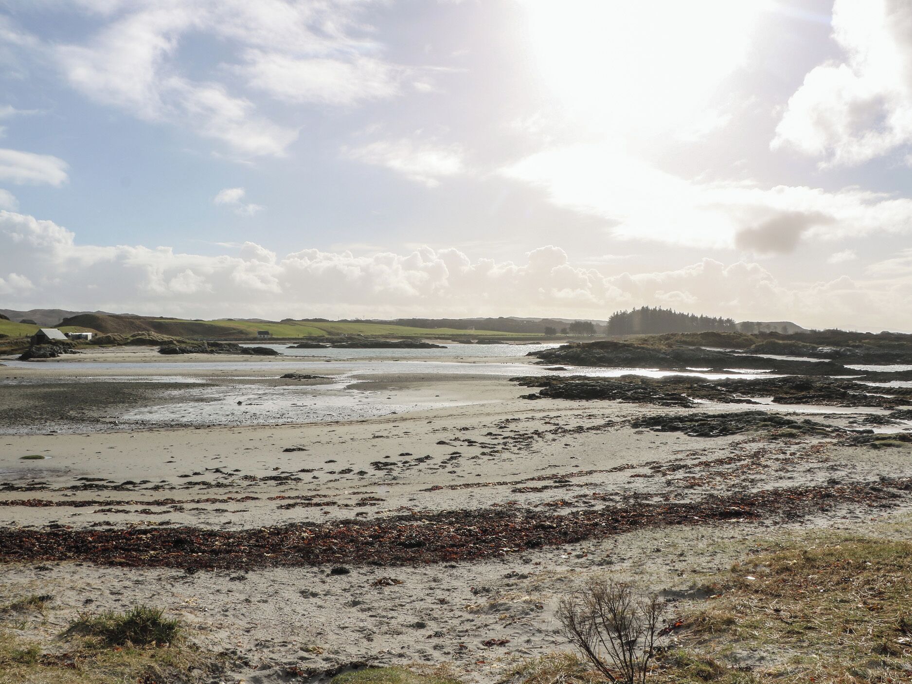A coastal landscape with a sandy beach, rocky outcrops, and a small building in the distance.