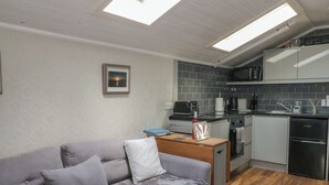 Ein kompakter Wohnbereich mit grauem Sofa, einem kleinen Holztisch, einer Küchenzeile mit modernen Geräten und einem Oberlicht über dem Sofa.