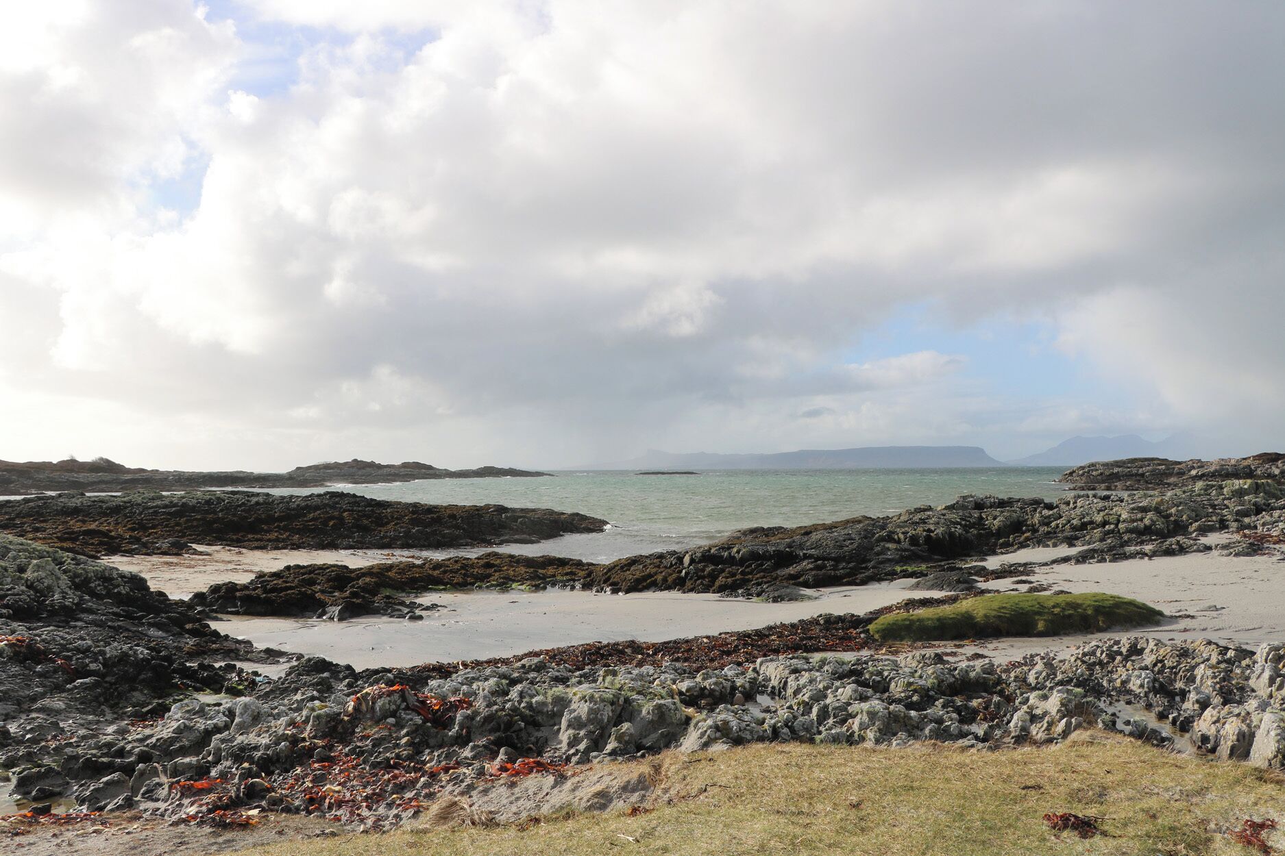 A rocky coastal landscape with a view of distant mountains under a cloudy sky.