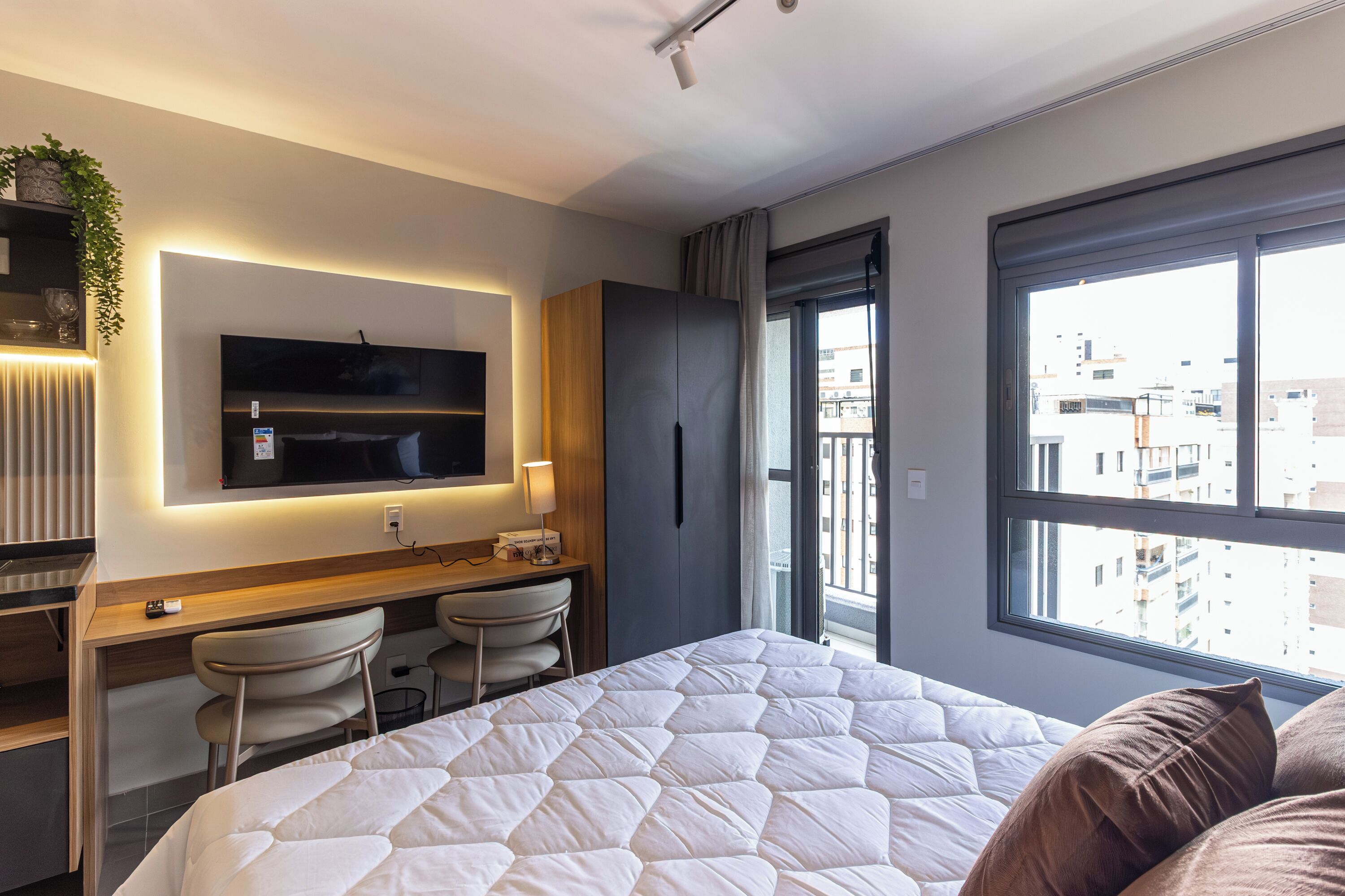 Una moderna camera d'hotel con un letto, una scrivania, sedie, una TV e un'ampia finestra con vista sulla città.