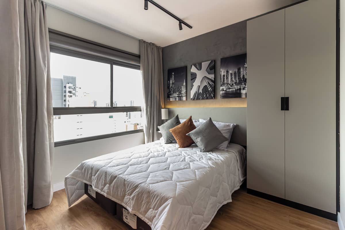 A modern bedroom with a large bed, cityscape artwork on the wall, and a view of buildings through the window.