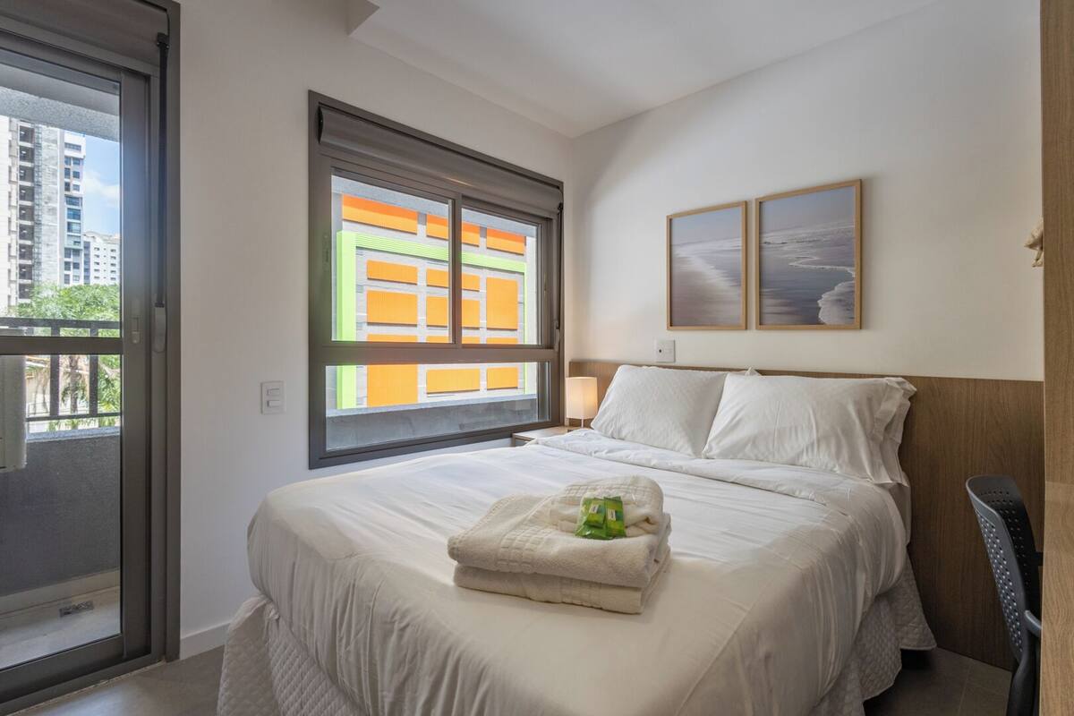 A hotel room with a bed, a window with a view of buildings, a nightstand with a lamp, and two framed pictures on the wall.