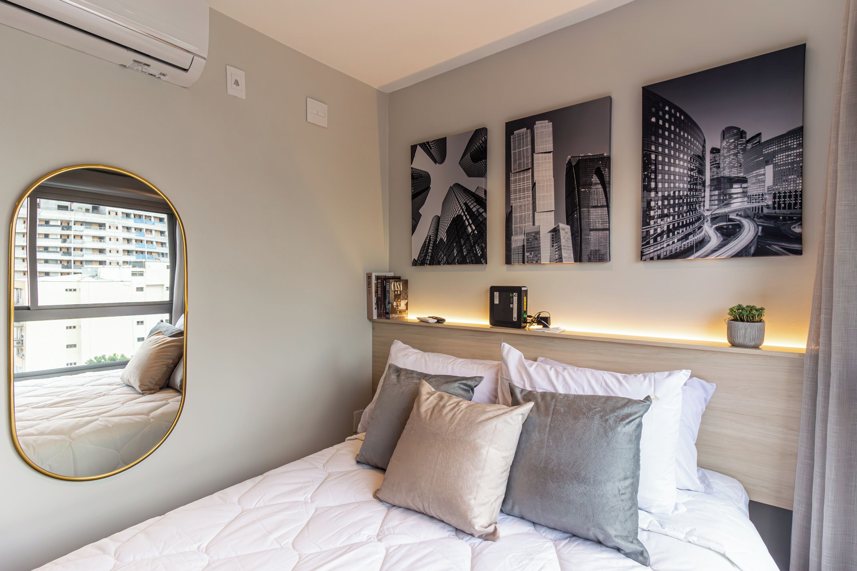 Una cama bien hecha con un cabecero, un espejo redondo y tres pinturas de paisajes urbanos en blanco y negro en la pared.