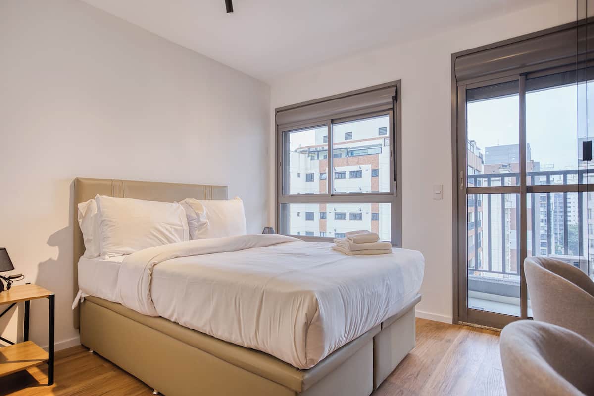 A modern bedroom with a large bed, a small bedside table, a chair, and a window with a city view.