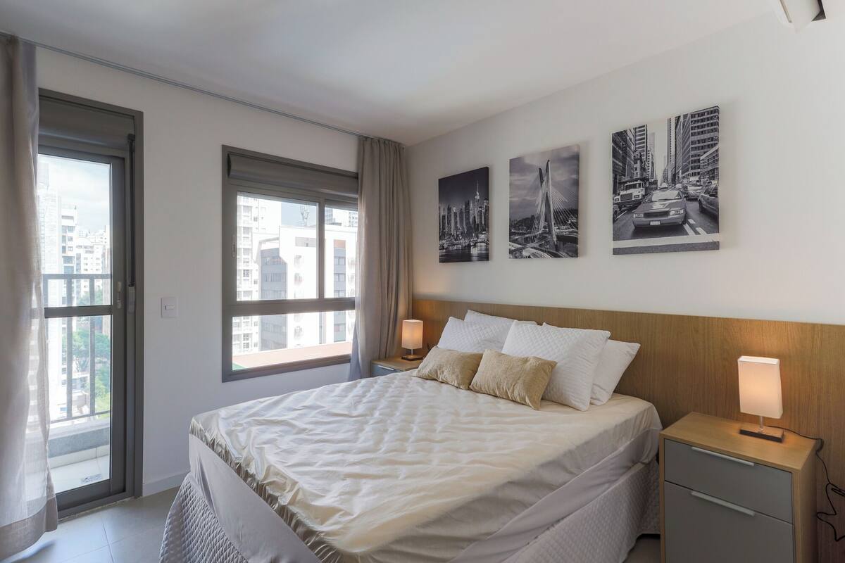 A hotel room with a large bed, two bedside tables, a window with curtains, and three black-and-white cityscape pictures on the wall.