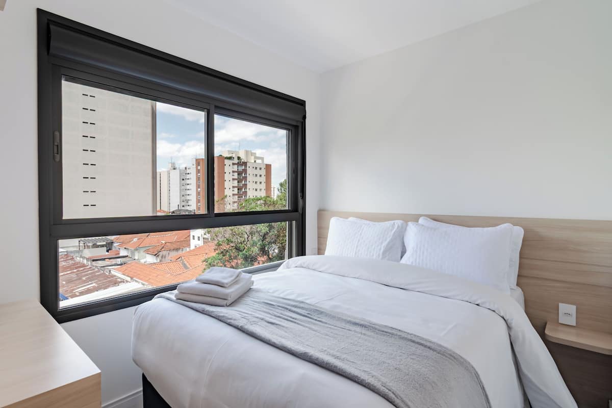 A hotel room with a large window, a neatly made bed, a bedside table, and a view of the cityscape.