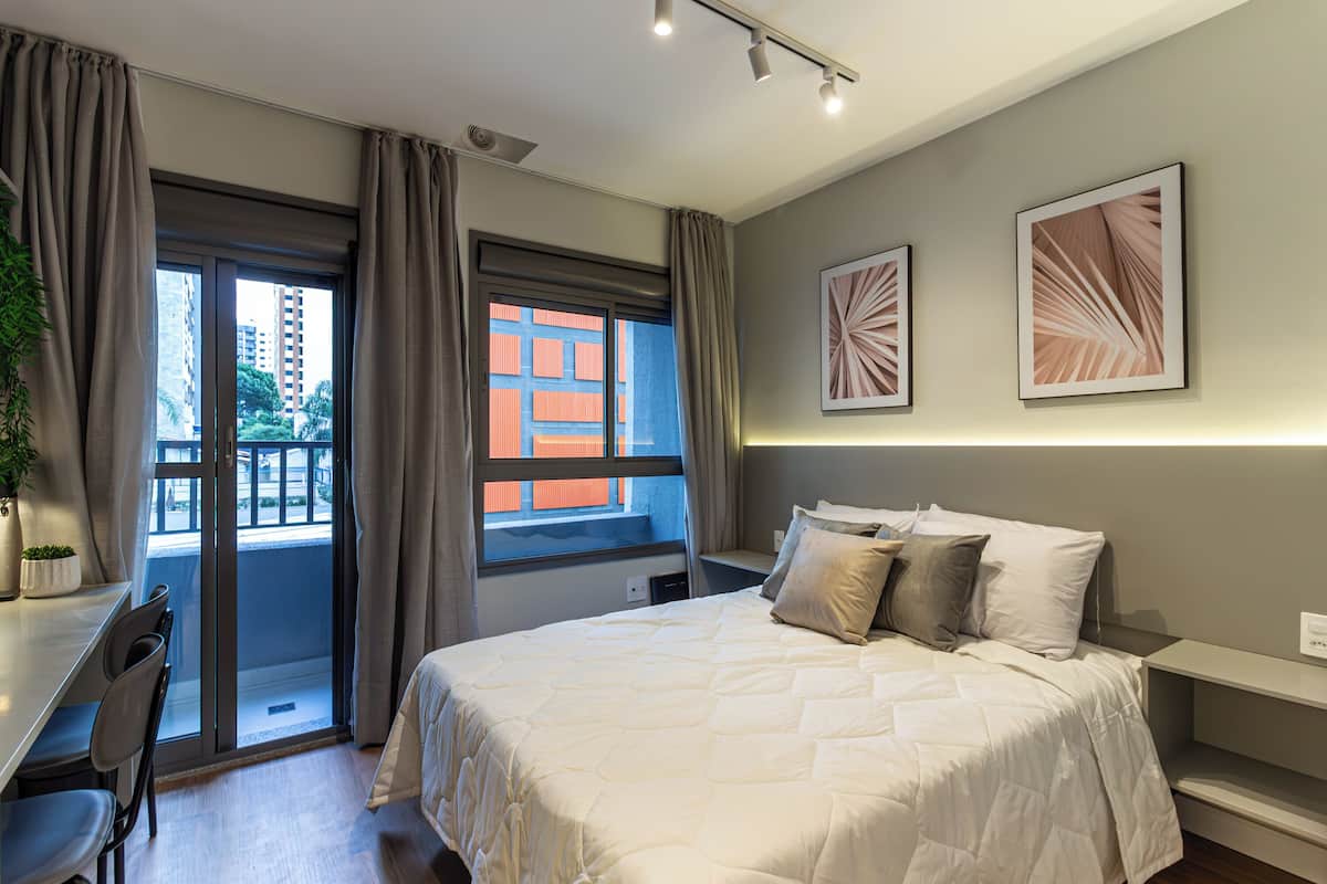 A hotel room with a bed, a desk, and a chair. There is a window with curtains, a plant, and artwork on the wall.