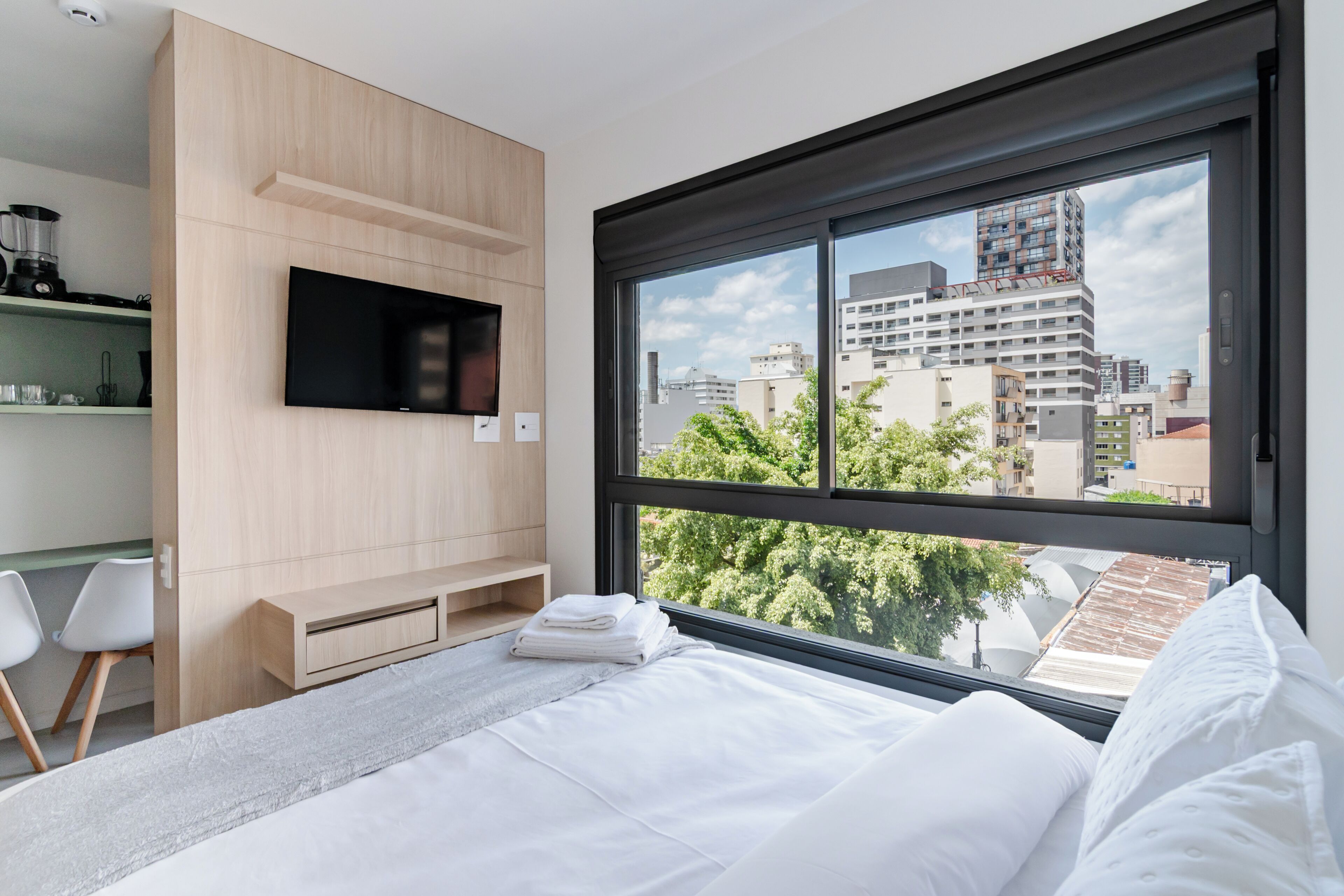 A modern hotel room with a bed, a TV mounted on the wall, a window with a city view, and a small seating area with chairs.
