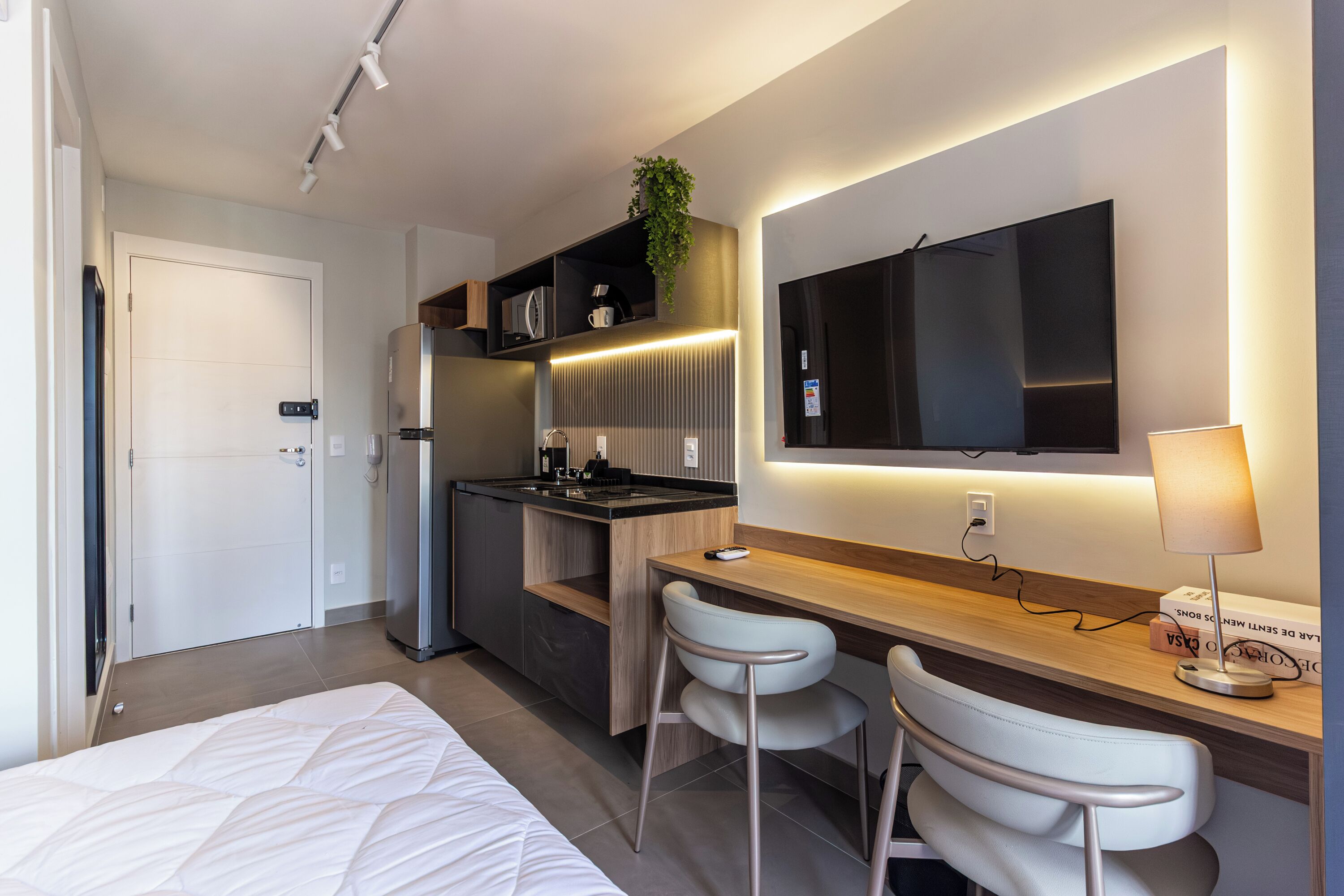A modern hotel room with a bed, a desk with two chairs, a kitchenette, a flat-screen TV, and a bedside lamp.