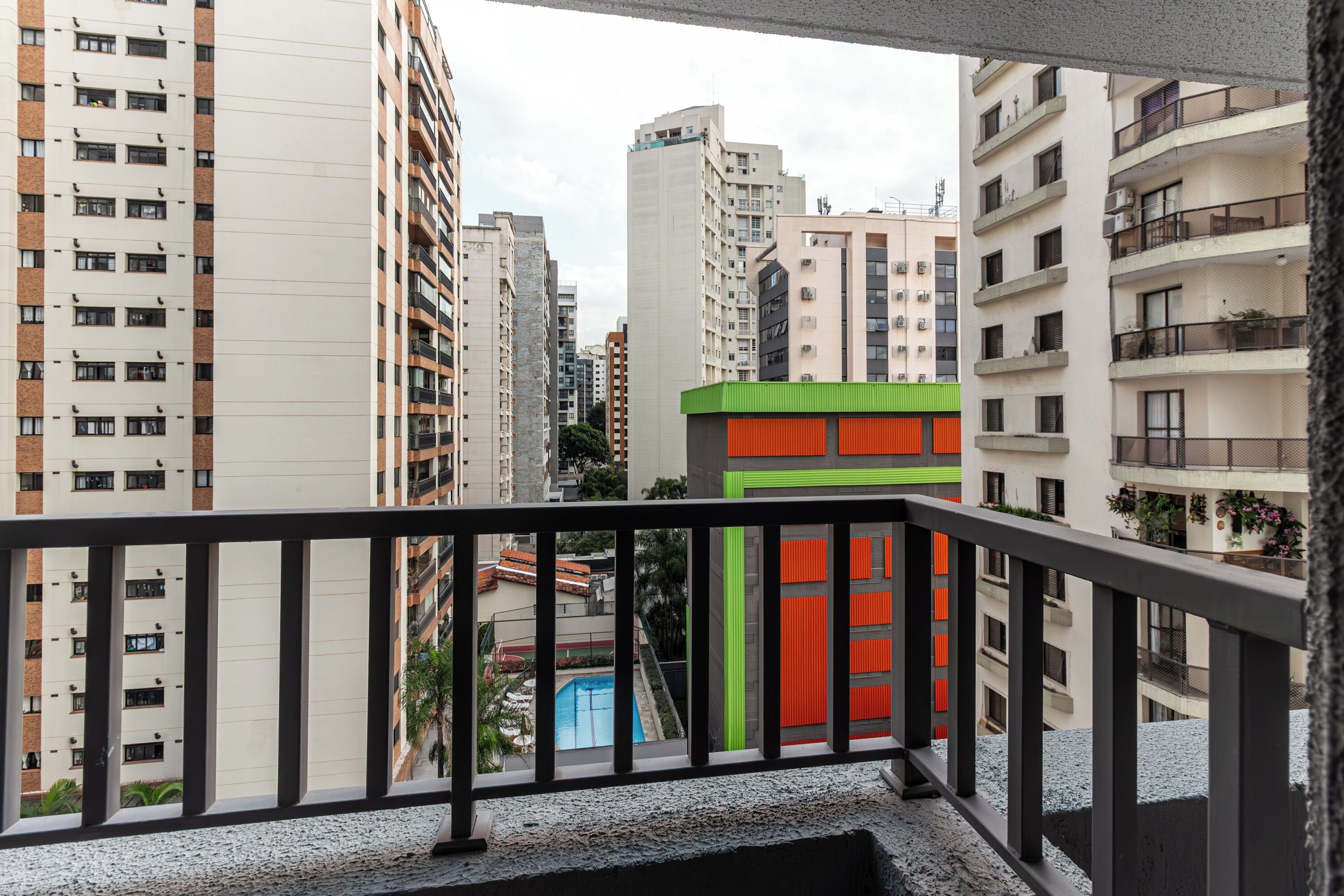 A balcony with a view of high-rise buildings, a swimming pool, and a green and orange building.