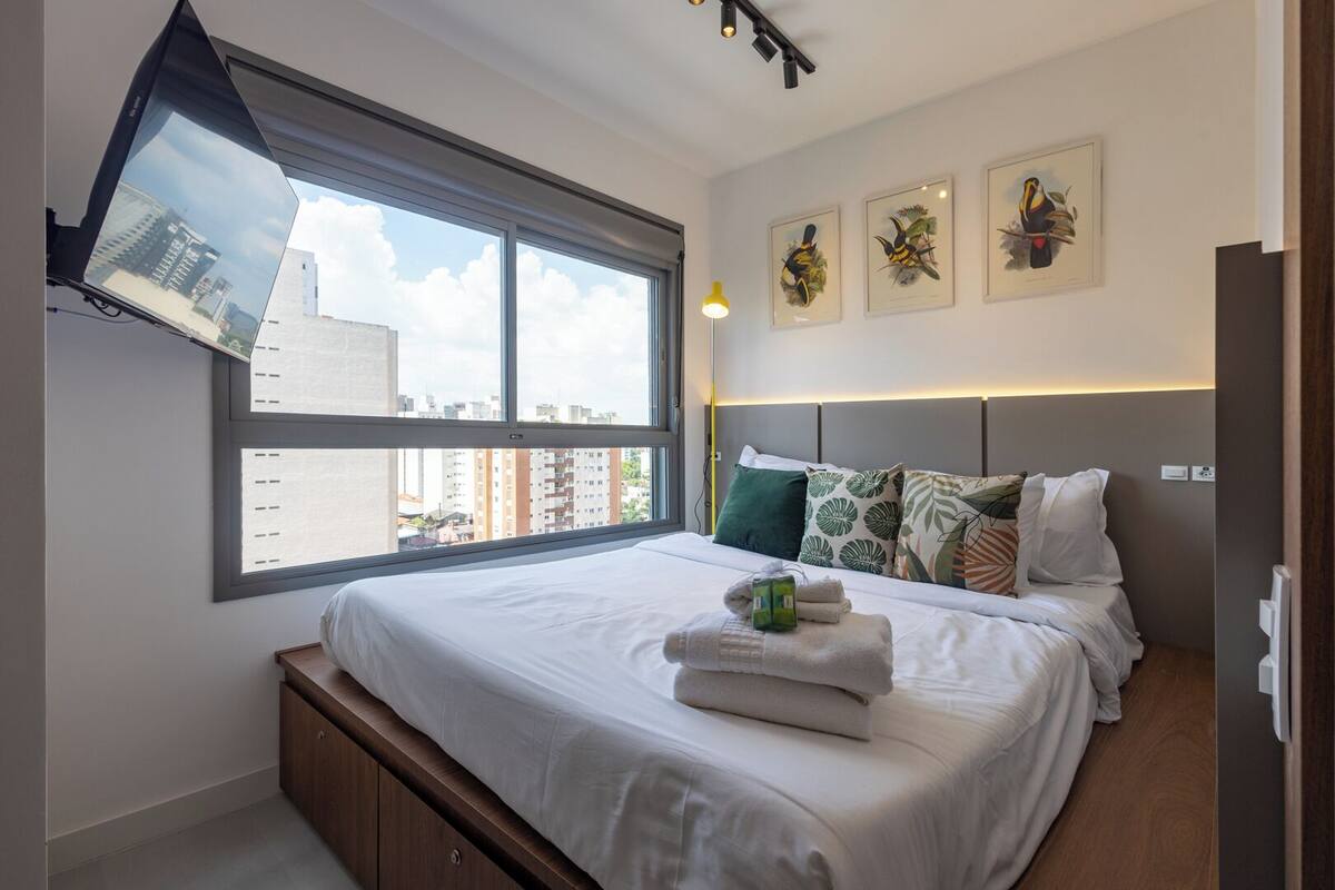 A modern bedroom with a large window, a bed with white linens, multiple pillows, and three framed bird paintings on the wall.