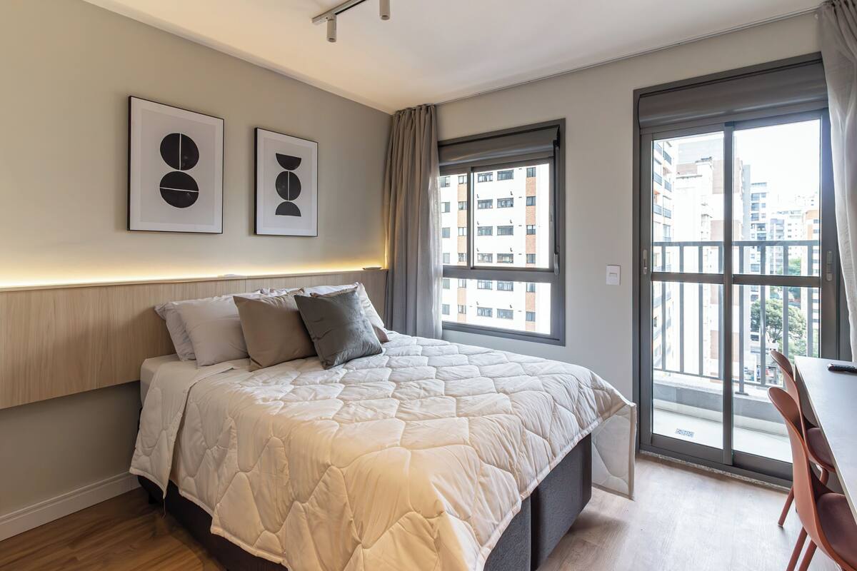 A modern bedroom with a large bed, a desk, and a view of the city through a large window.
