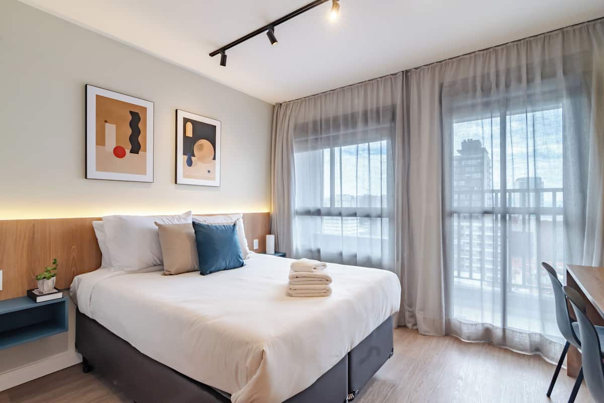 A modern hotel room with a large bed, a desk, and a view of the city through sheer curtains.