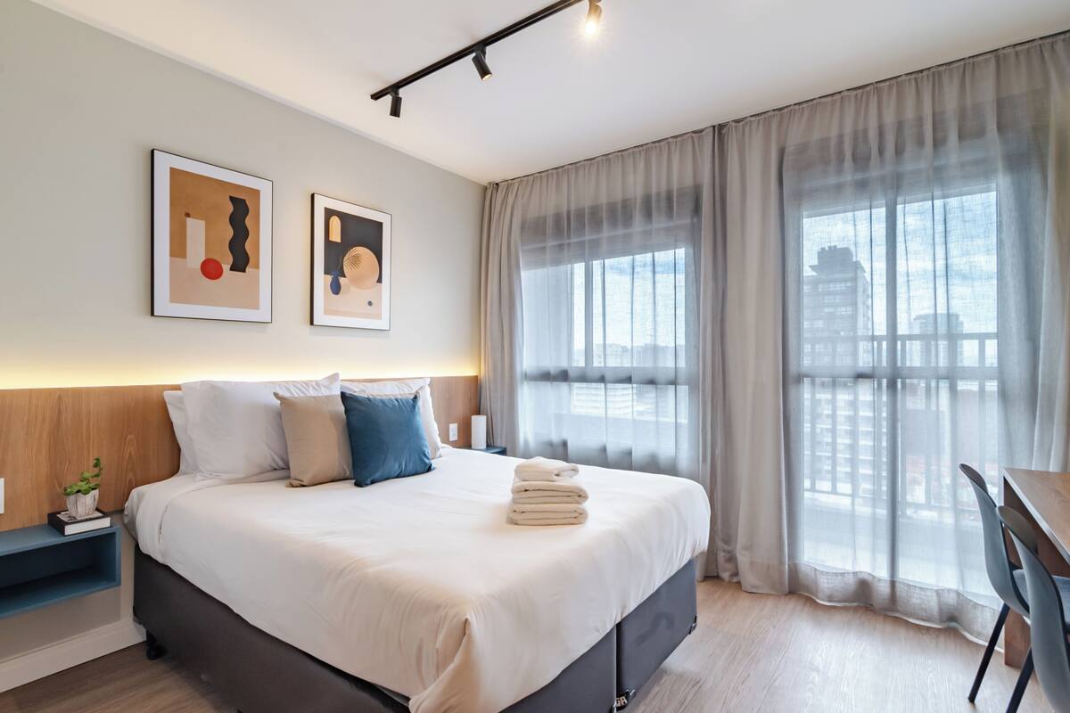 A modern hotel room with a large bed, a desk, and a view of the city through sheer curtains.