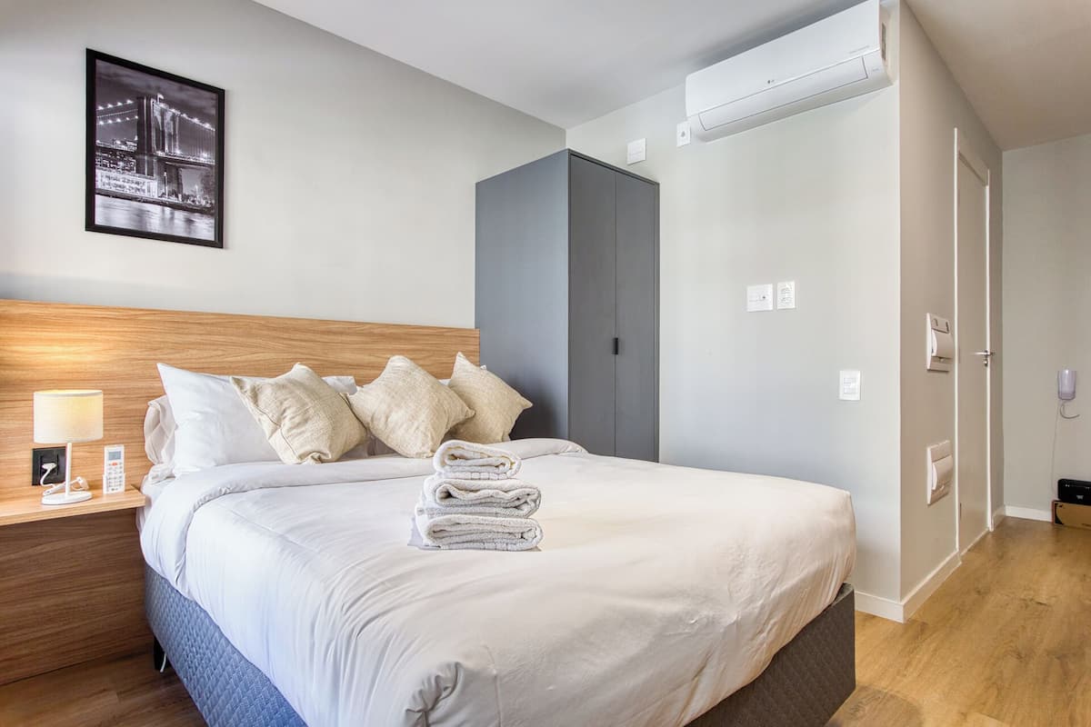 A hotel room with a bed, a nightstand, a lamp, a folded towel, a framed picture on the wall, a wardrobe, and an air conditioning unit.