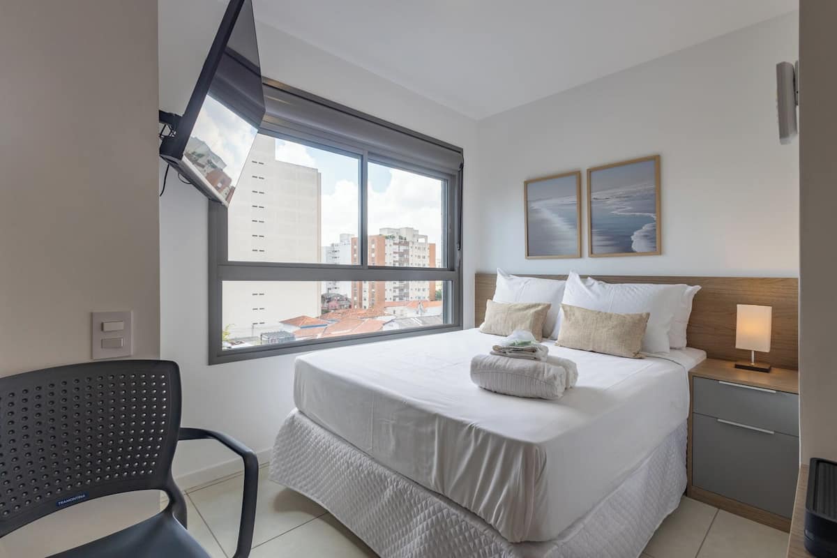 A hotel room with a large bed, a chair, a nightstand, and a window with a city view.