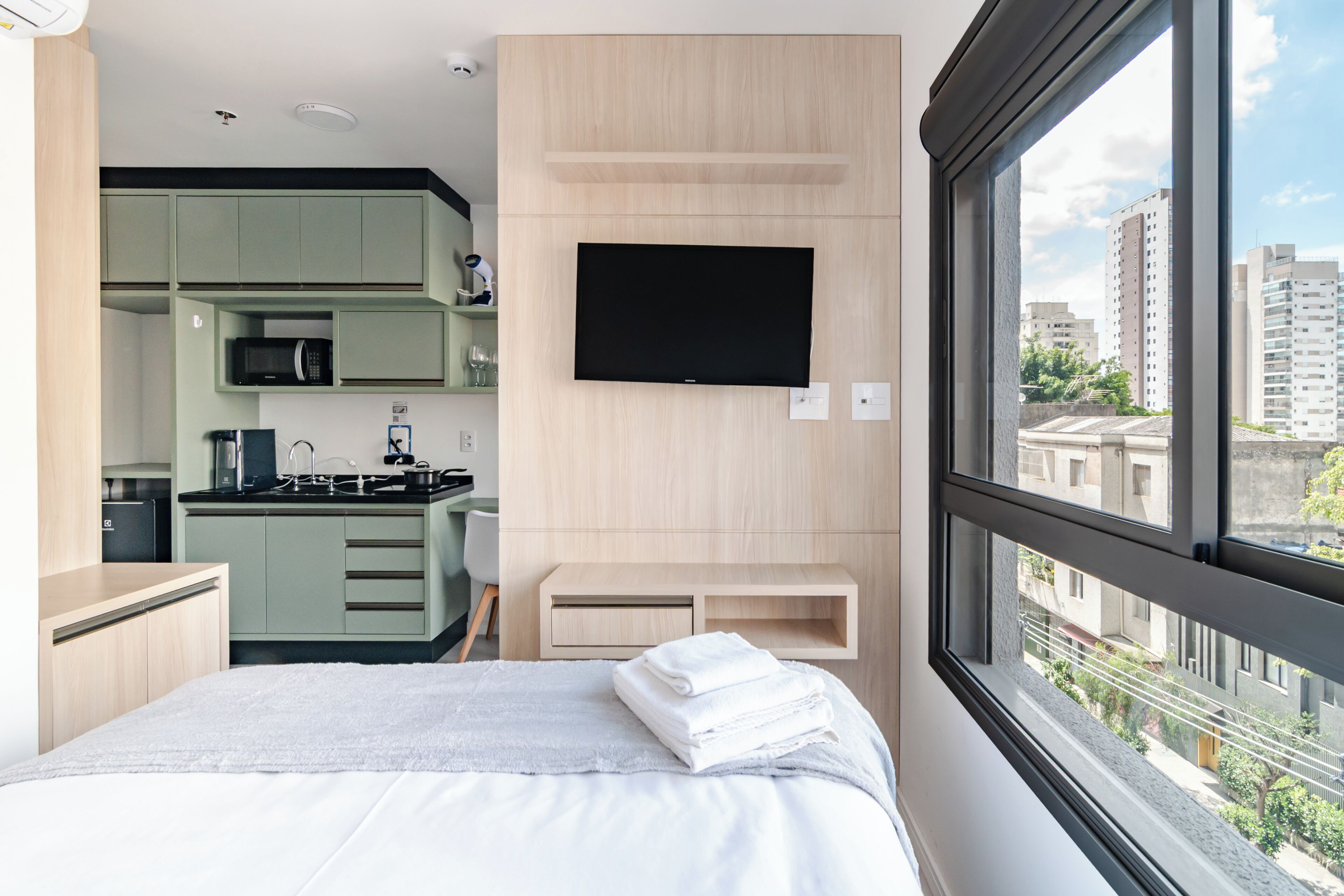 Een moderne hotelkamer met een bed, een aan de muur bevestigde televisie, een kitchenette en uitzicht op de stad.