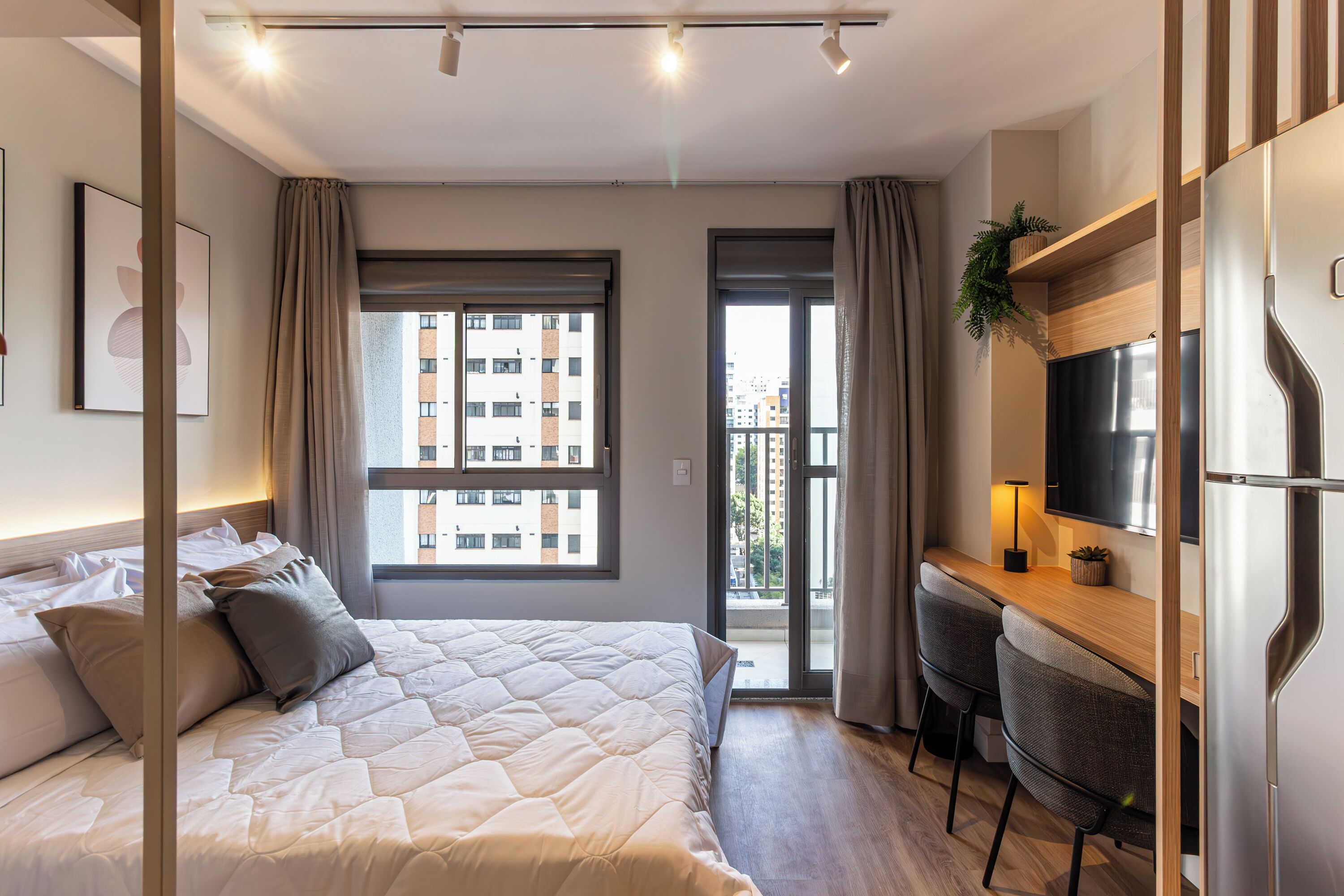A modern hotel room with a large bed, a desk with two chairs, a flat-screen TV, and a balcony with a view of the city.
