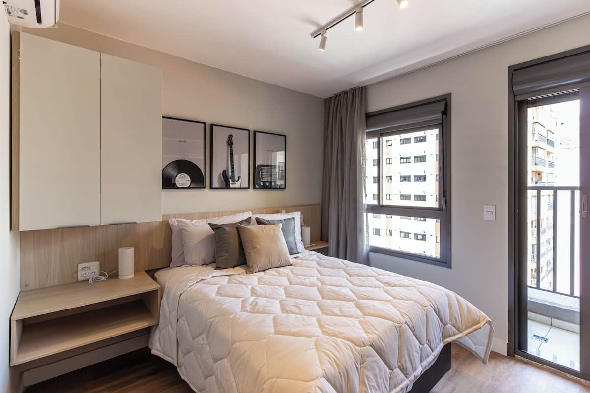 A modern bedroom with a large bed, a bedside table, a wall-mounted shelf, and a window with a view of buildings.