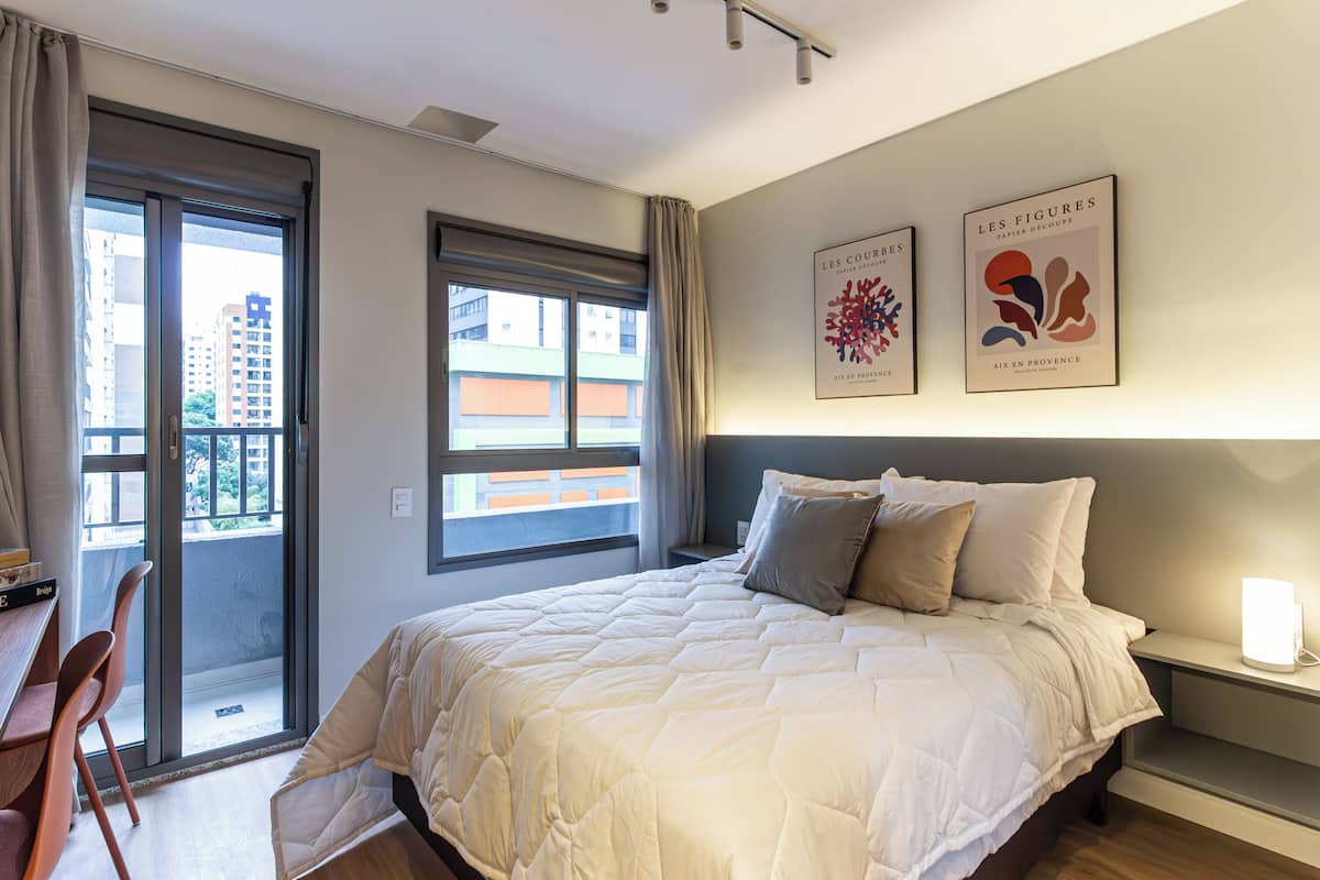 A hotel room with a bed, a desk, a chair, a window with curtains, and two wall-mounted artworks.
