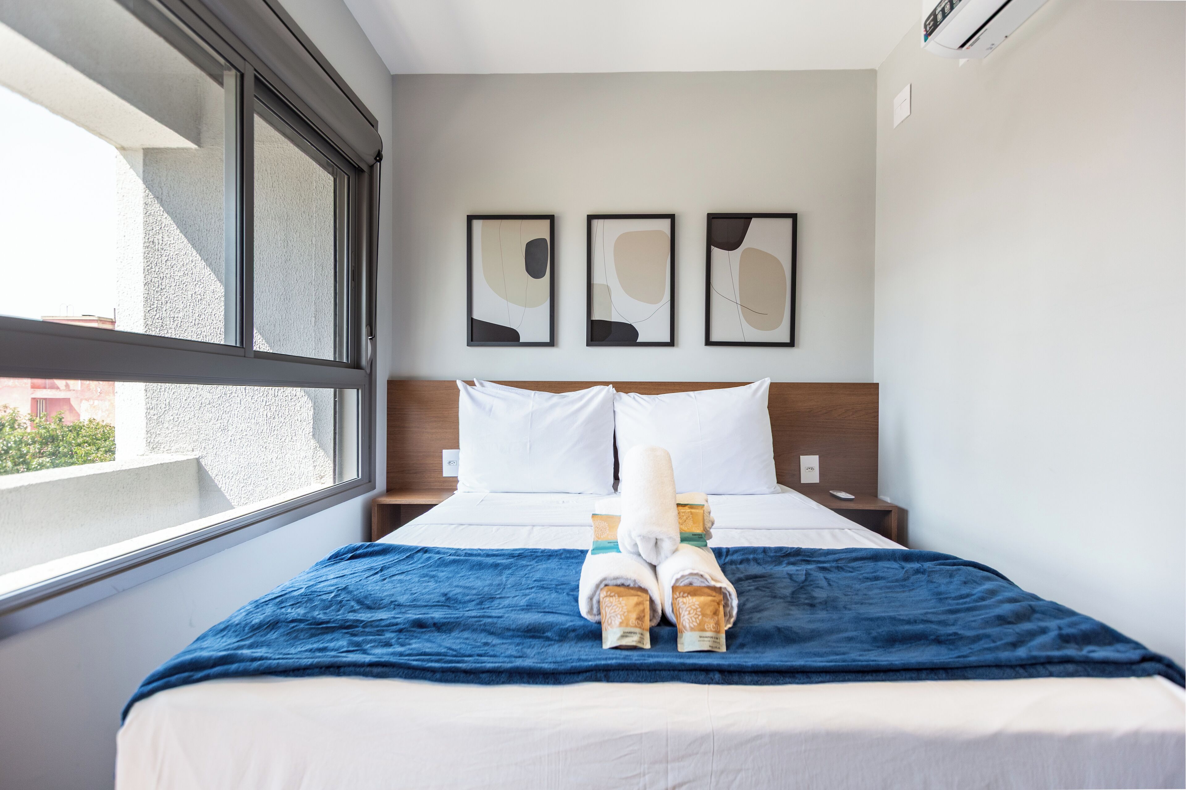 Ein Hotelzimmer mit großem Fenster, einem Bett mit blauer Decke, weißen Kissen und zwei gerahmten abstrakten Kunstwerken an der Wand.