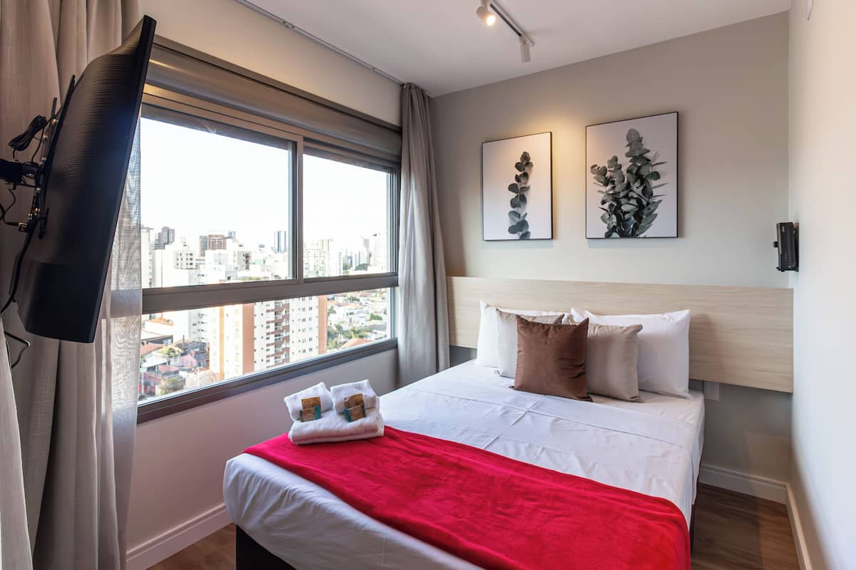 A hotel room with a bed, a window with curtains, a flat-screen TV, and artwork on the wall.