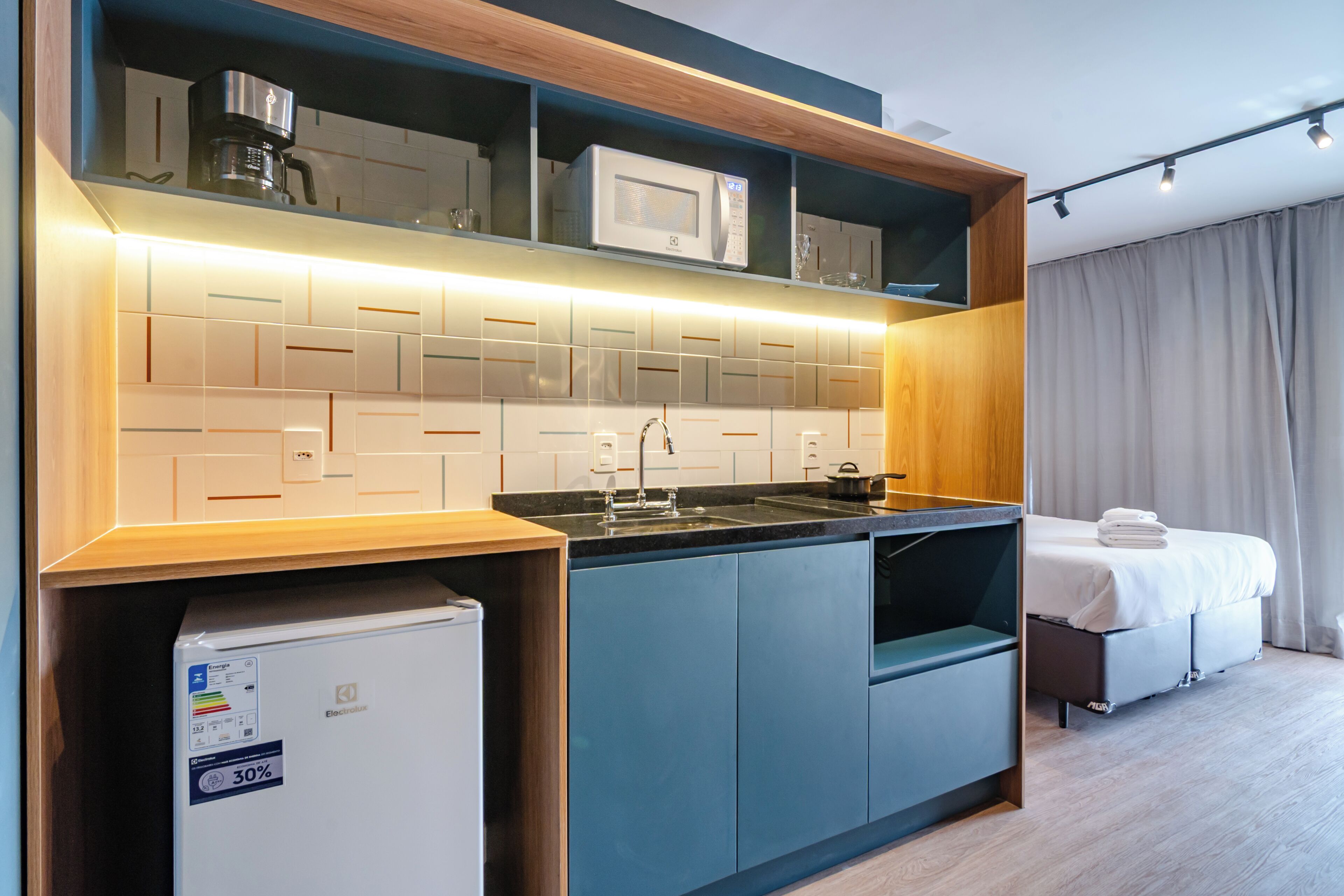 Habitación de hotel compacta con kitchenette, una cama y un ventanal con cortinas.