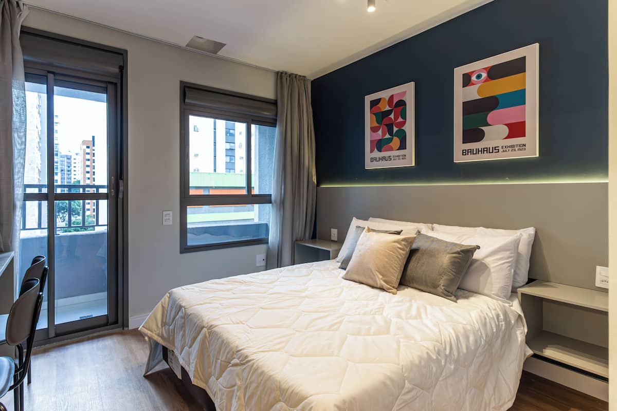 A hotel room with a large bed, a desk, a chair, a window with a view of buildings, and two artworks on the wall.