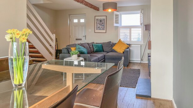 A modern living room with a glass dining table, a grey sofa with colorful cushions, a staircase with exposed wooden beams, and a large window with blinds. - 3 bedroom accommodation in Monyash, near Bakewell (Monyash, near Bakewell)