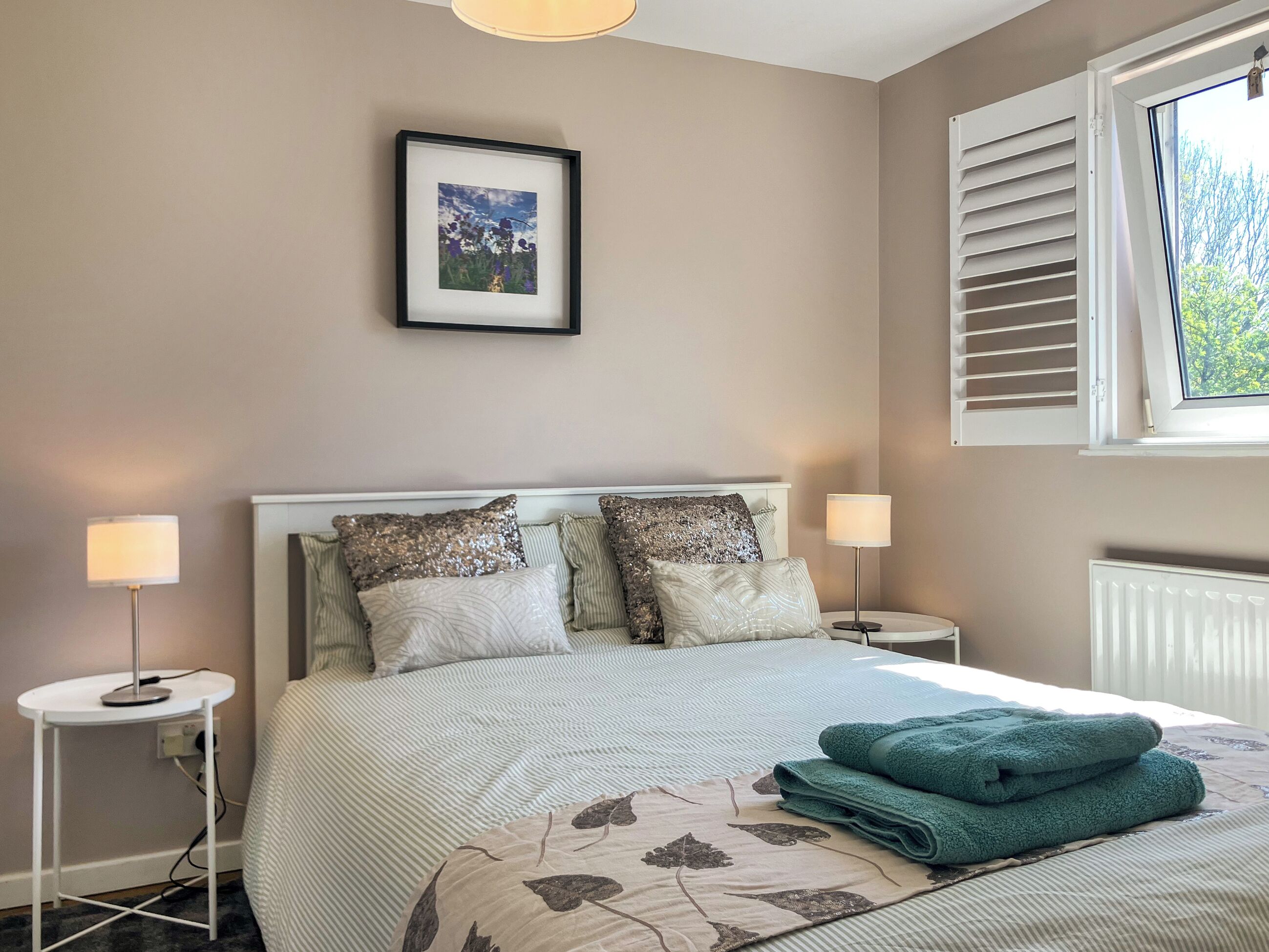 Une chambre à coucher bien rangée, avec un lit, deux lampes de chevet, un tableau encadré au mur et une fenêtre avec des volets.