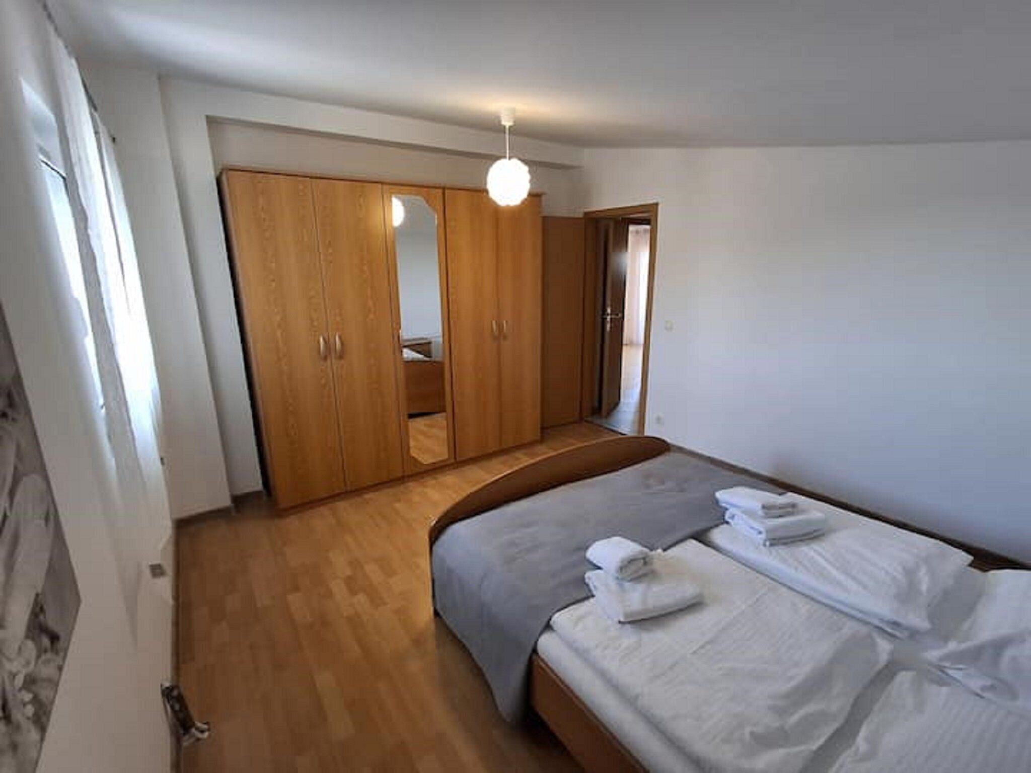 A bedroom with a wooden wardrobe, a bed with white linens, and a small bathroom visible through a door.