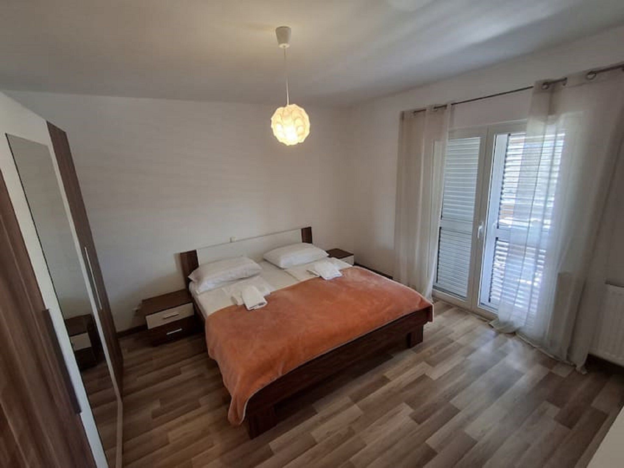 A bedroom with a wooden bed, a nightstand, a hanging light fixture, and a view of the outside through a window with blinds.