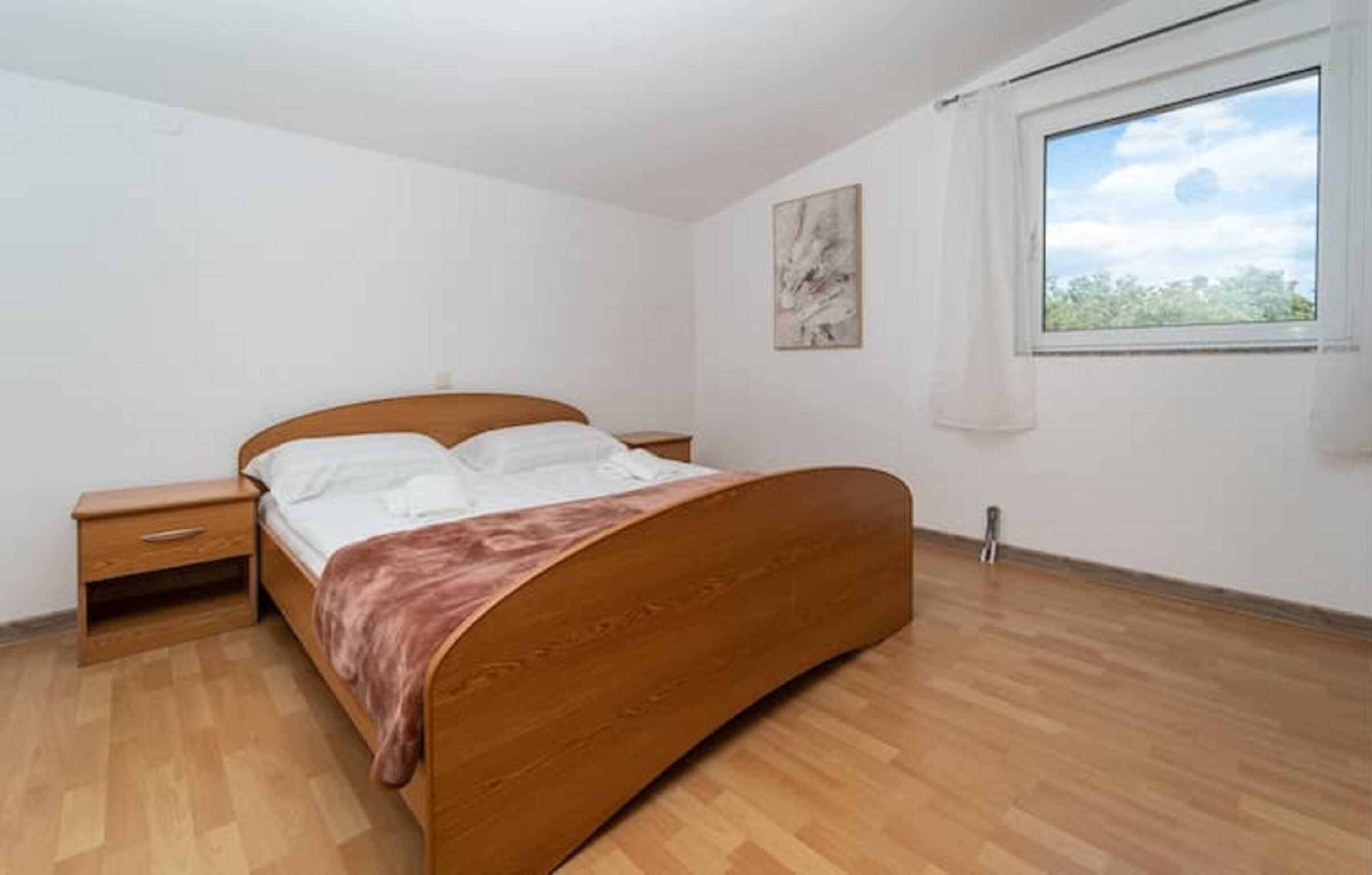 Une chambre avec un lit en bois, une table de chevet, une fenêtre donnant sur un paysage verdoyant et un tableau accroché au mur.